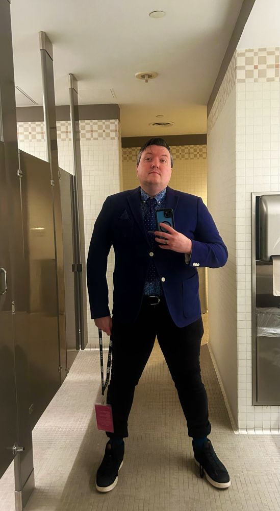 A picture of a man in front of full length mirror in a restroom. He’s wearing a blue blazer, black pants, black and white sneakers, a floral dress shirt and a black tie with blue spots.