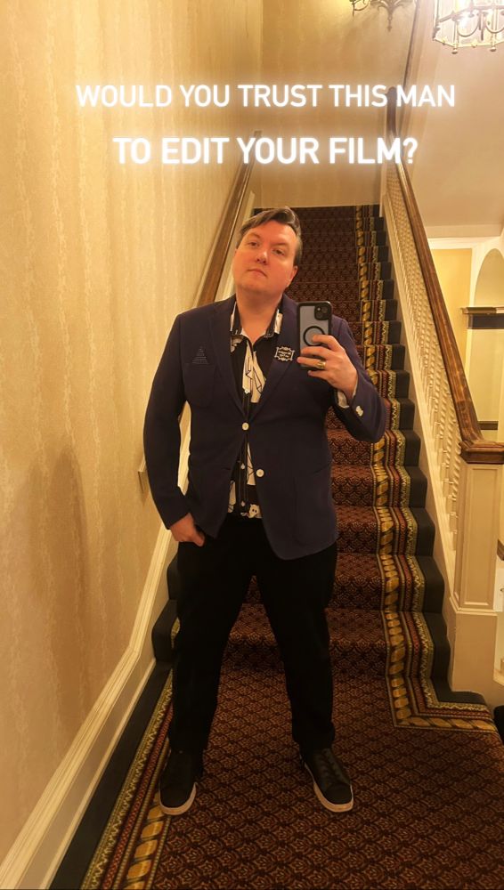 A picture of me, a queer man, a mirror selfie, wearing a blazer and a button up shirt with ghosts over it and a pin that says all vampires are gay.