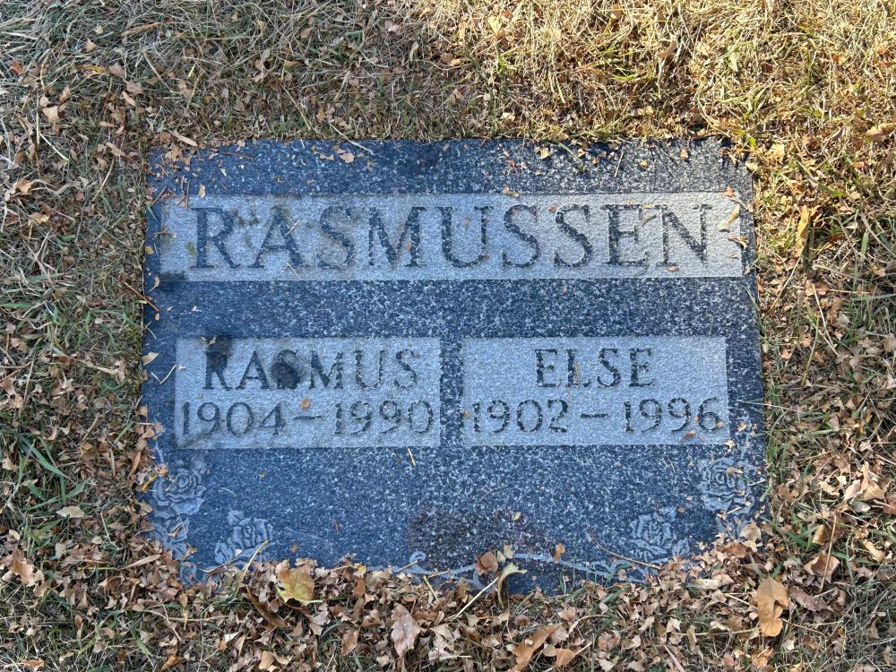 Photo of a flat cemetery marker of dark stone surrounded by autumn grass. The family name is Rasmussen, the deceased Rasmus and Else. Both passed in the 1990s. Engravings of roses on the bottom corners.