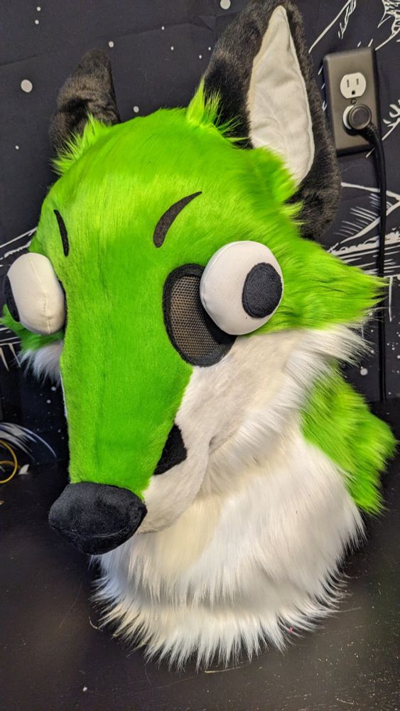 A very silly and stupid-looking fox fursuit head with green fur, buggy eyes, and a little pink tongue poking out to the side. His ears are flopped back and his eyes are pointed in different directions. 