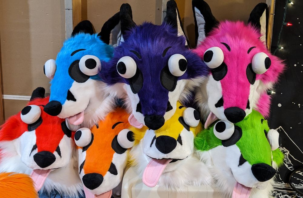 A stack of rainbow colored dumb fox fursuit heads. Top row - blue, purple, and pink. Bottom row - red, orange, yellow, and green. They all have big buggy eyes and pink tongues hanging out of their mouths. So silly. Very fox. Wow. 