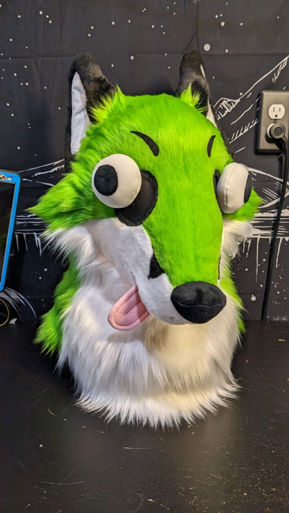 A very silly and stupid-looking fox fursuit head with green fur, buggy eyes, and a little pink tongue poking out to the side. His ears are flopped back and his eyes are pointed in different directions. 