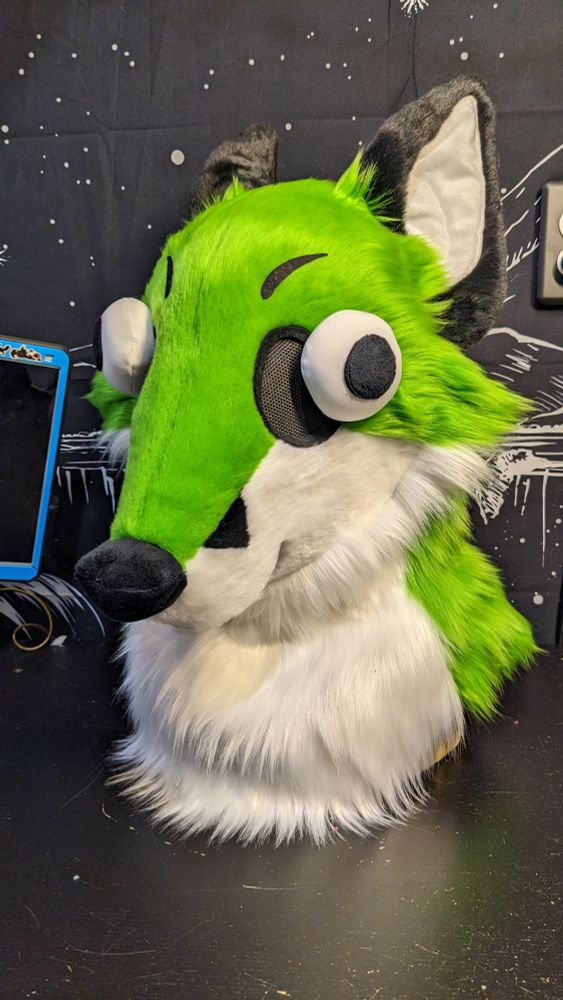 A very silly and stupid-looking fox fursuit head with green fur, buggy eyes, and a little pink tongue poking out to the side. His ears are flopped back and his eyes are pointed in different directions. 