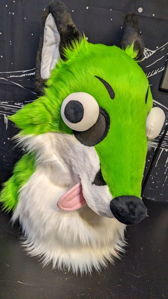 A very silly and stupid-looking fox fursuit head with green fur, buggy eyes, and a little pink tongue poking out to the side. His ears are flopped back and his eyes are pointed in different directions. 