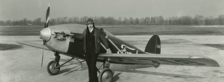 a man in leather helmet and pilot goggles standing in front of the Ford Flivver, a single-seat airplane bearing the Ford motor company script logo, probably in around 1927. The Flivver was, historian wits like to say, easy to fly; more difficult, alas, to land.