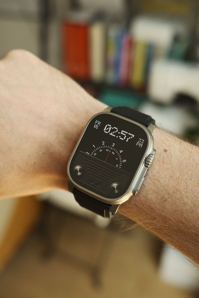 A close-up image of a person's wrist wearing a modern smartwatch with a rectangular face and a black strap. The watch face design features a unique, dark, retro-inspired graphic resembling an old radio dial with a needle indicator and two round knobs at the bottom. The background is softly blurred, highlighting the watch and wrist in sharp focus.