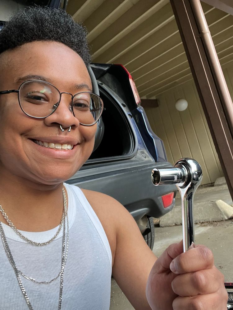 a photo of skye holding a socket wrench. he’s wearing a white tank-top, headphones, and three necklaces of varying lengths. they’re smiling in the photo as they hold up the socket wrench to give the viewer a sense they know what they’re doing, even though they absolutely don’t. skye’s car, gina, is in the background with her trunk open.