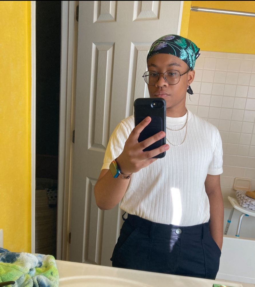 skye takes a terrible bathroom mirror selfie. they’re wearing a white ribbed shirt and navy blue pants, two necklaces one is shorter than the other, a flowery bandana, and the hand holding his phone has an apple watch with a rainbow cover. in the background, you can see a shower chair with blocking pads stacked on top of it inside a bathtub. the door behind skye is open, revealing a just barely visible hamper in their dark room.