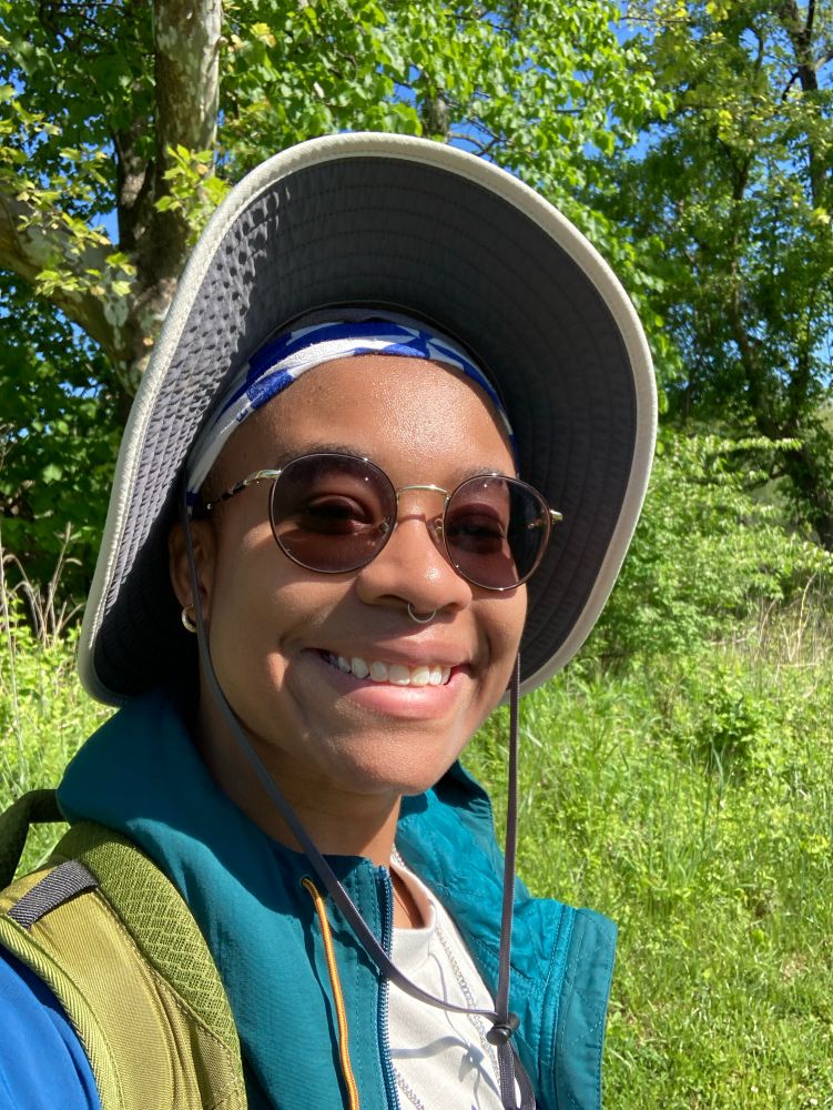 skye is outside in the field. they’re wearing a sunhat with a bandana underneath, a dark teal coat, and a yellow-green backpack. he’s smiling in the image, and their glasses are slightly tinted from the sun.