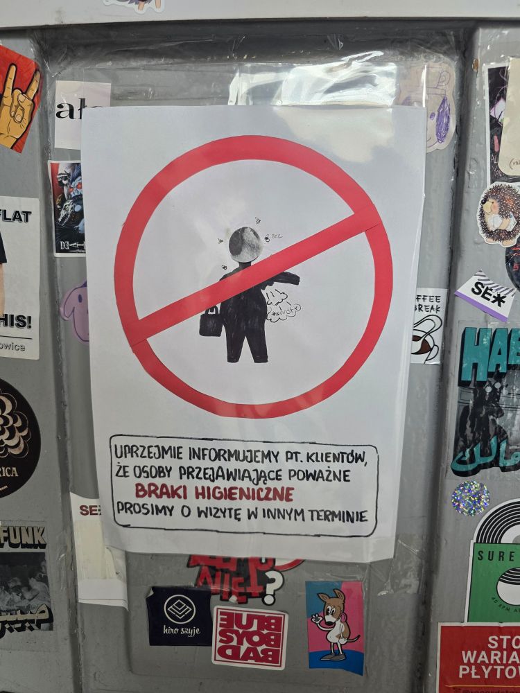 A close-up photograph of a white paper sign attached to a textured surface. The sign features a red circle with a diagonal line (a "no" symbol) over a black stick figure holding a briefcase in one hand and a crumpled white item in the other. Below the image is Polish text in black font, stating "We kindly inform our customers that people with serious hygiene deficiencies are asked to visit at another time." The sign is surrounded by various stickers, including a yellow hand giving a thumbs-up, a sticker with the word "FLAT," another with "HIS!," "FFEE BREAK," "SEX," "FUNK," "SURE," and others with illegible text.