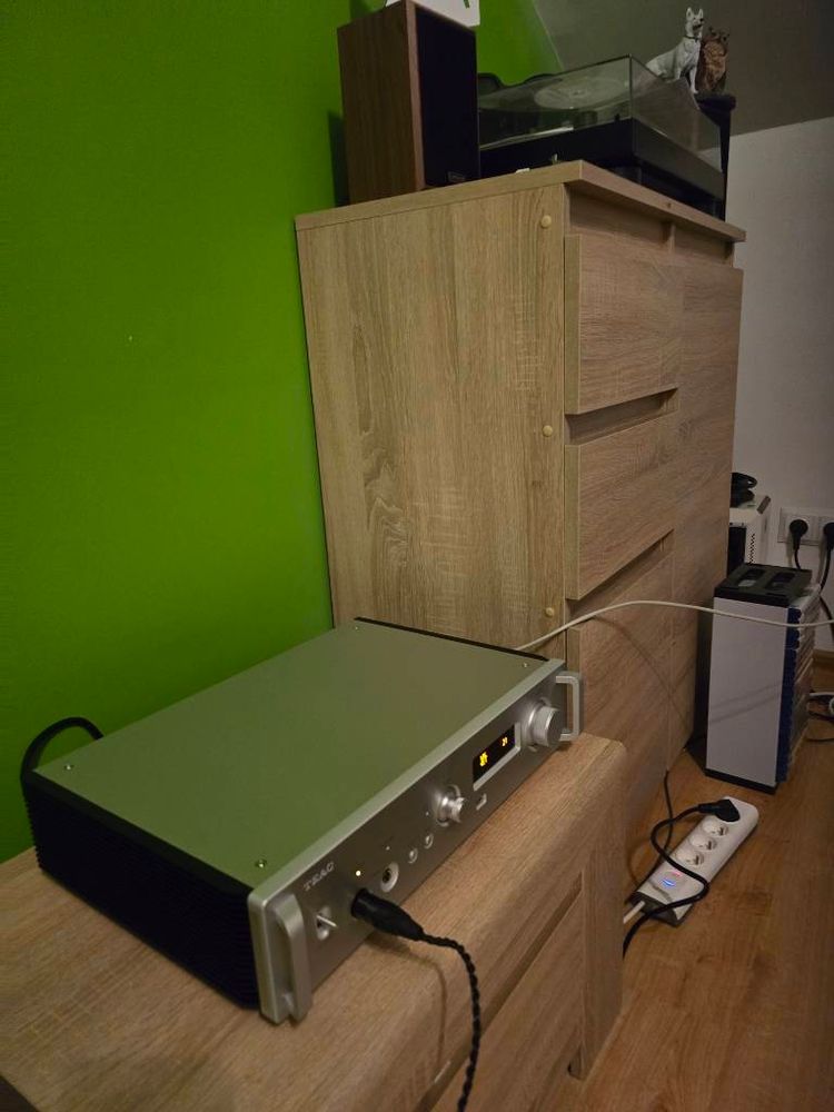 The image shows an audio amplifier or receiver with a silver/gray front panel sitting on top of a light wood cabinet or media console. The amplifier has various control knobs and buttons on its front panel, with some small display lights visible. The cabinet has multiple drawers with recessed handles. Both are positioned against a bright green wall. There appears to be other electronic equipment visible in the background, including what might be a power strip or surge protector on the floor. The setting appears to be a home entertainment or audio setup.