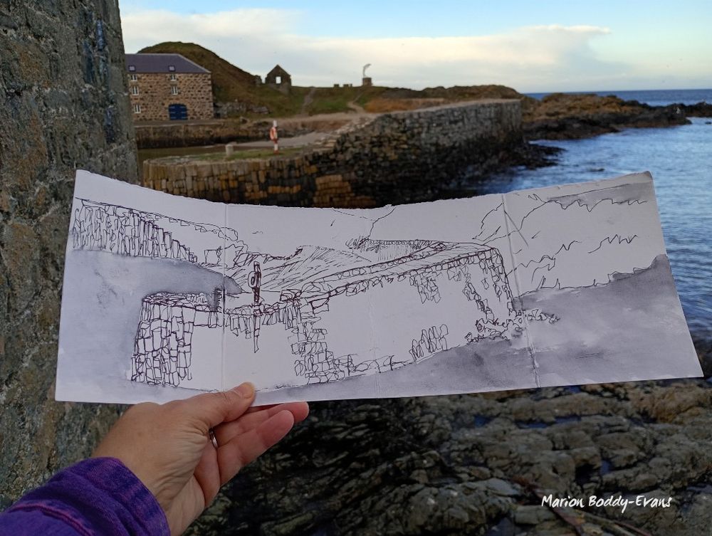 My hand holding my ink drawing of the old harbour at Portsoy, Scotland, with the scene visible behind it. To my left is the stone wall of a building.