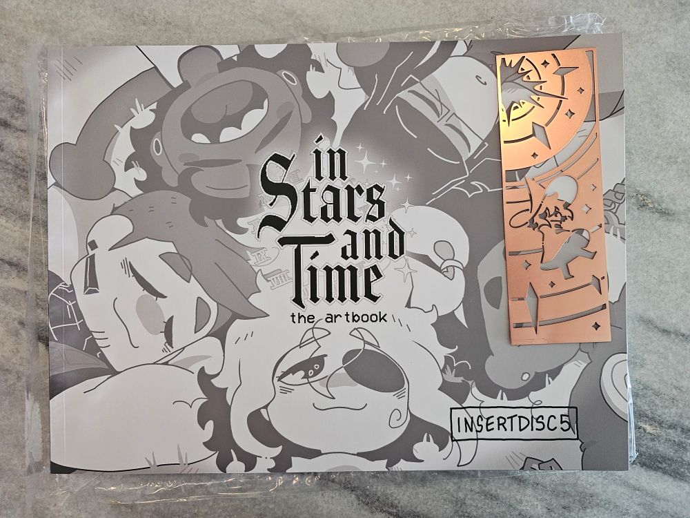 The In Stars and Time artbook and a metal bookmark with a siffrin design
