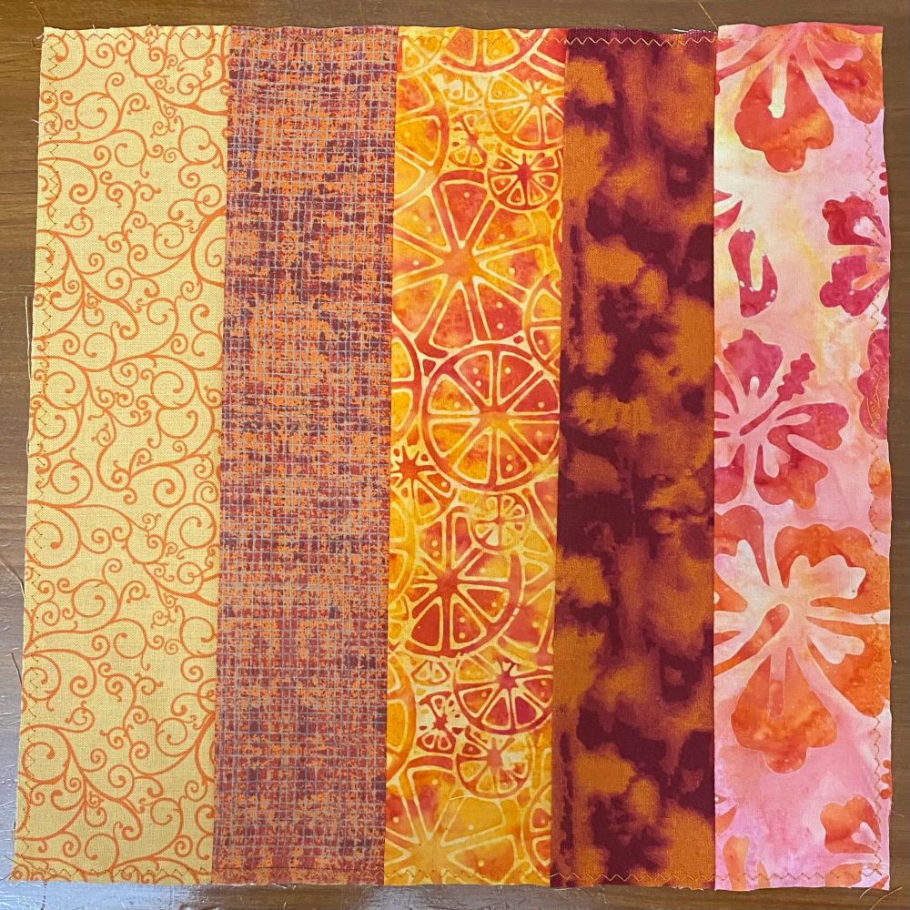 A “rail fence” quilt block in predominantly orange, red, and gold colors. The block has five different fabrics, three of which have abstract patterns, though one depicts orange slices and another flowers.