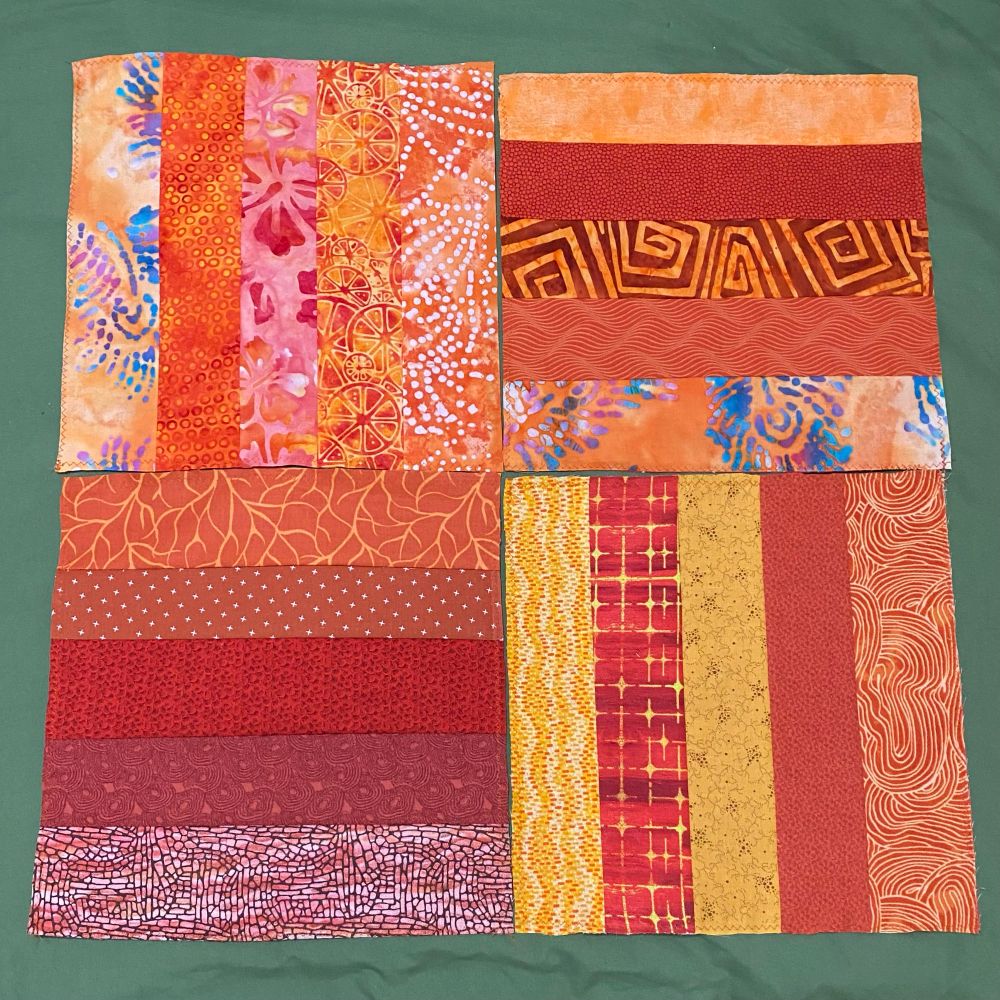 Multiple “rail fence” quilt blocks in predominantly orange, red, and gold colors. Most blocks have five different fabric strips, many of which have abstract patterns, though some depict animals, fruit, flames, leaves, stars & planets, or flowers.