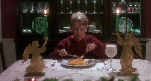 Scene from Home Alone where Kevin McAllister (played by actor Macaulay Culkin) is sitting at his family's dining table, set with a white tablecloth and angel shaped candleholders lifting green, lit candles. He's holding up a knife and fork to dig in to his plate of max and cheese. There's a wine glass (specifically a burgundy glass) of milk to compliment his meal.