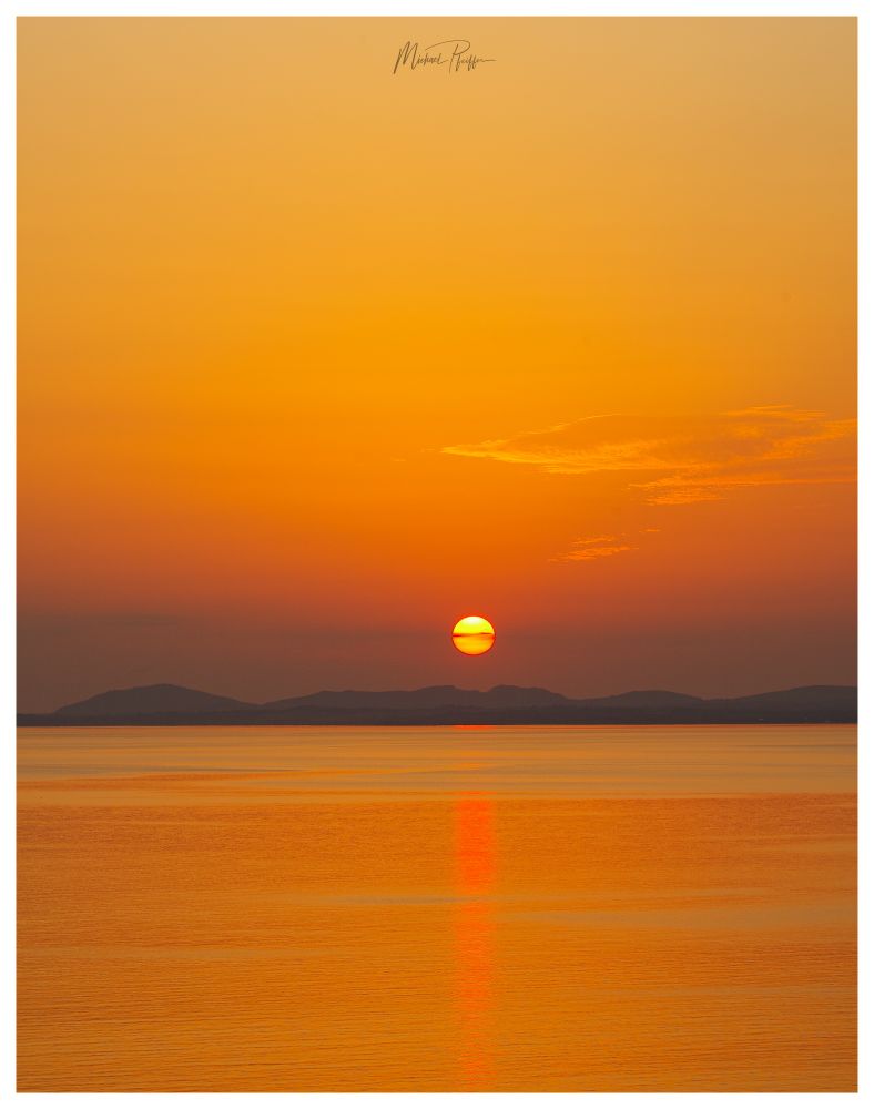 Farbenfroher Sonnenuntergang über dem Meer und einigen Bergen in der Ferne.Die Sonne in der Mitte des Bildes nur knapp über dem Horizont und herangezoomt