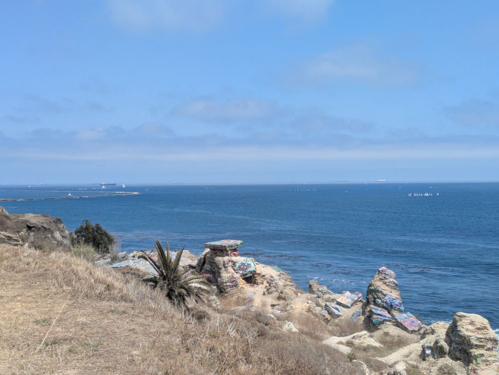 A photo of sunken city along the Pacific coast. Out on the water there are multiple groups of tiny sailboats in the midst of a race. The sky is blue and dotted with clouds on the horizon and it's a bright, sunny day.