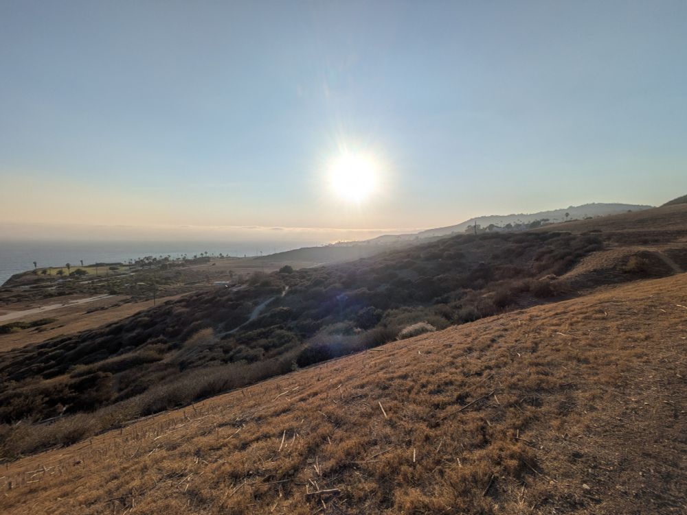 A photograph of some hills and gullies covered in brown grasses and some shrubs that fall off towards the Pacific on the left side of the photo. The sun is dead center, just above the horizon in a clear blue sky with a marine layer of clouds underneath.