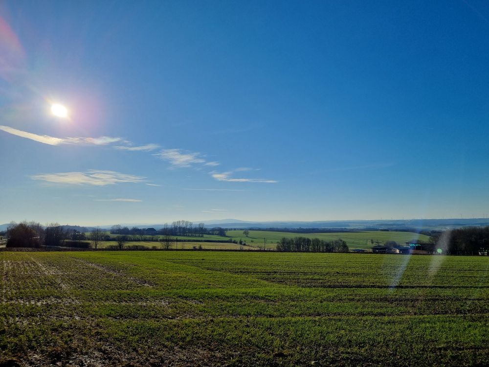 Landschaftsfoto mit blauem Himmel, Sonne und kleinen Wolken.