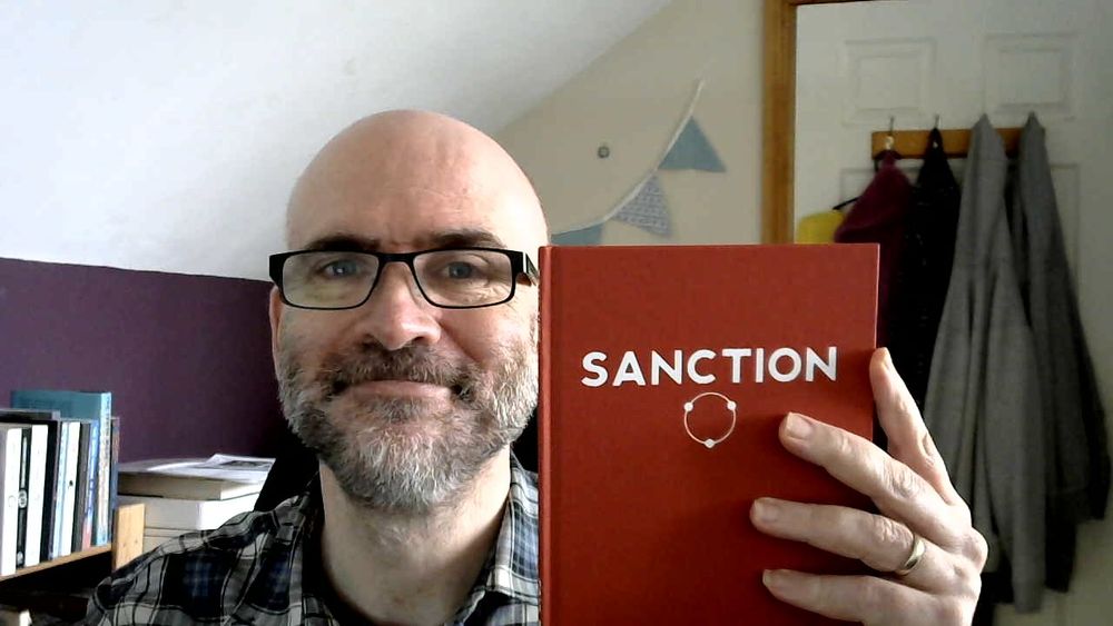 I am holding up a copy of the roleplaying rule set 'SANCTION. It's a hardback, red, with Sanction in white upper case letters and below that a white circle with three dots spaced round the circumference.