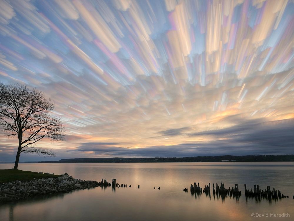 Long exposure of clouds moving across a bay at sunrise with a section of shoreline and a row of old wooden pilings in the foreground 