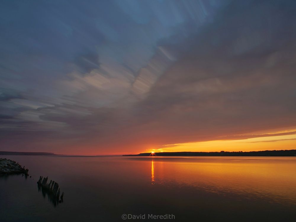 Colpoy's Bay, Ontario at sunrise with the sun appearing over the Niagara Escarpment on the east side of the bay. There is a band of cloud diagonally across the sky and old wooden pilings sticking out of the water in the foreground. There is a band of yellow sky between the band of cloud and the Niagara Escarpment. 