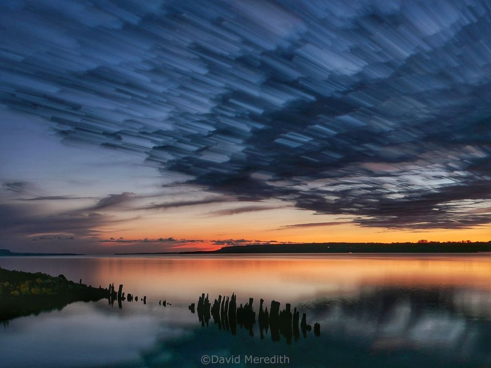 Long exposure of cloud streaking across Colpoy's Bay shortly before sunrise with a section of shoreline and a row of old wooden piles sticking out of the water in the foreground 