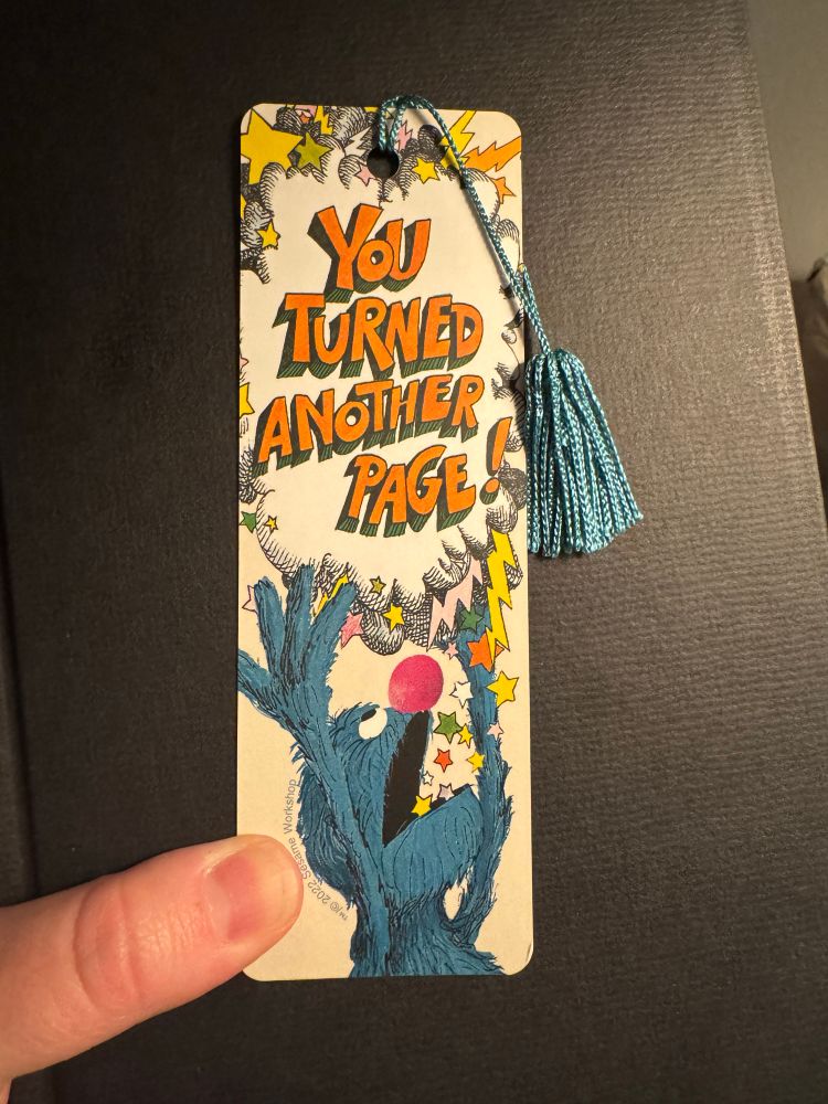 A bookmark on a black book. The bookmark has an illustration of Grover the muppet, from the book “The Monster At The End of This Book.” Grover has thrown up his hands, completely exasperated, with a speech bubble saying “YOU TURNED ANOTHER PAGE!”