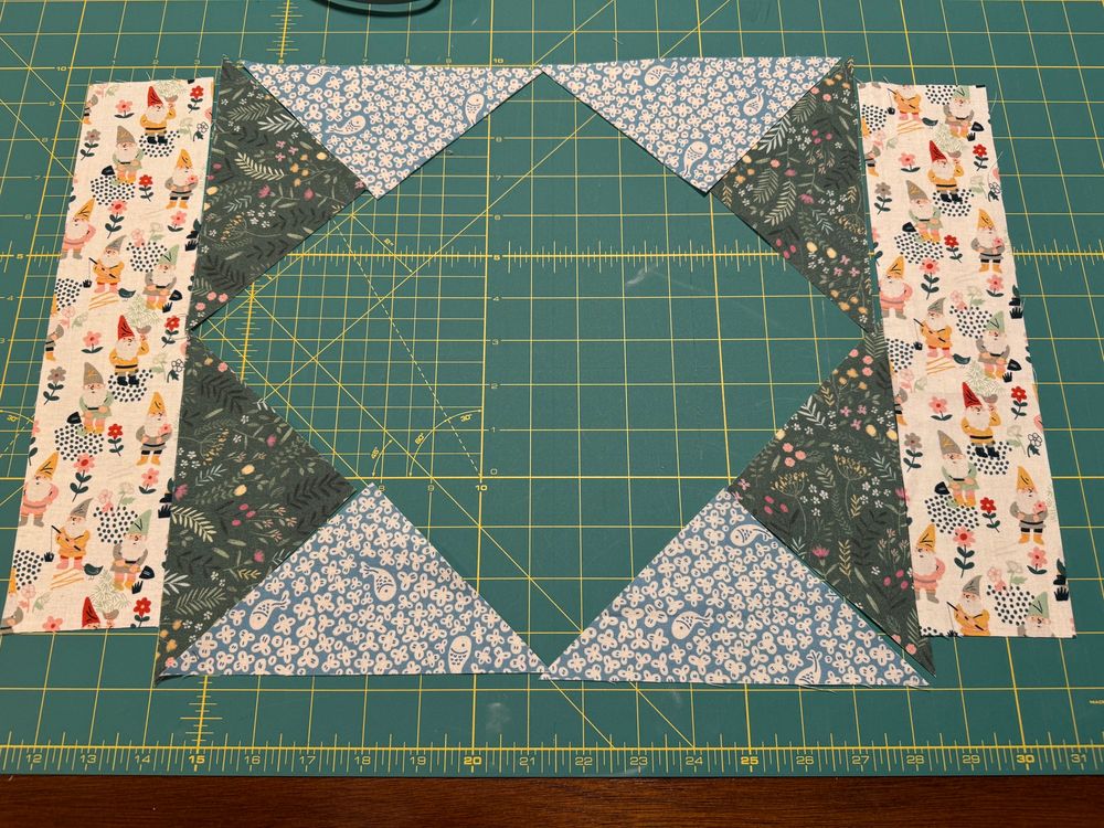 The fabric for a placemat before I sew them all together. The long thin strips of fabric on either side have adorable gnomes on them. The interior of the place mat has an open spot for a square turned 90 degrees (so the corners at at the poles). Around the square are alternating triangles of green fabric with wild flowers and blue fabric with fish and flowers. 