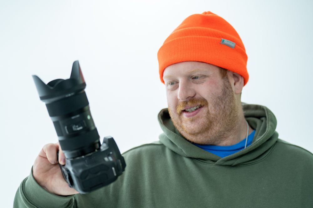 Man in orange beanie cap and hoodie holding a cannon camera with a large lens standing in front of a white cyclorama wall. 