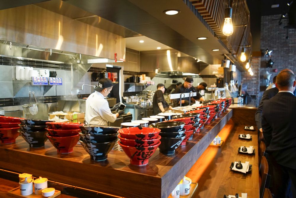 Red and black ramen bowls in foreground at Jinya Ramen Bar grand opening in New Jersey. The kitchen is buzzing with activity before the evening rush, and everything is in order. Aperture mode shooting a Medium depth of field focusing on the bowls in the foreground  and the chef in middle and focus blurs as we follow the line of bowls down the line and see rest of kitchen staff blurred in the back. 