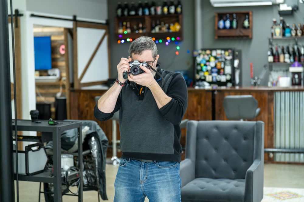 Film photographer in grey vest, standing in ambassador Studios New Jersey photography studio, looking through a canon camera view finder, capturing an image of another photographer. 