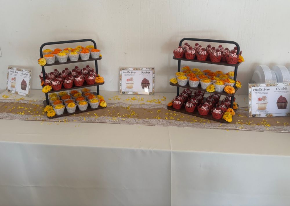 A zoomed out photo showing two of the 3-tiered shelves beside one another on the same table. The left one has shelves of the flower cupcakes on the top and bottom, with the red cupcakes in the middle. The right one is inverted, with two shelves of the red cupcakes and one of the flowery ones. Three plastic signs sit upright on the table, showing a diagram of the two differen types of cupcakes. The flower ones are vanilla bean, while the red ones are chocolate. The yellow and orange flower petals are scattered all around the table. 