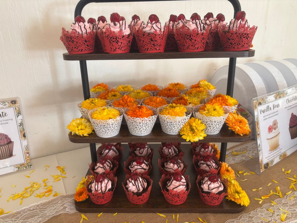 A 3-tiered shelf display sits on a white table in front of a white wall. Each tier is a 1 foot by 1 foot square of wood, and holds several rows of cupcakes. The top and bottom shelves contain brown cupcakes with pink frosting, a lacy red cupcake holder, and a cherry on top. The middle shelf holds light cupcakes with a lacy white cupcake holder, and the tops dominated by 1 or 2 flowers. The flowers are various shades of orange and yellow, with dozens of long narrow petals fanned around the center. Several more flowers are placed in between cupcakes on the shelves of the display, and loose petals are scattered on the rustic beige table runner beneath them. 