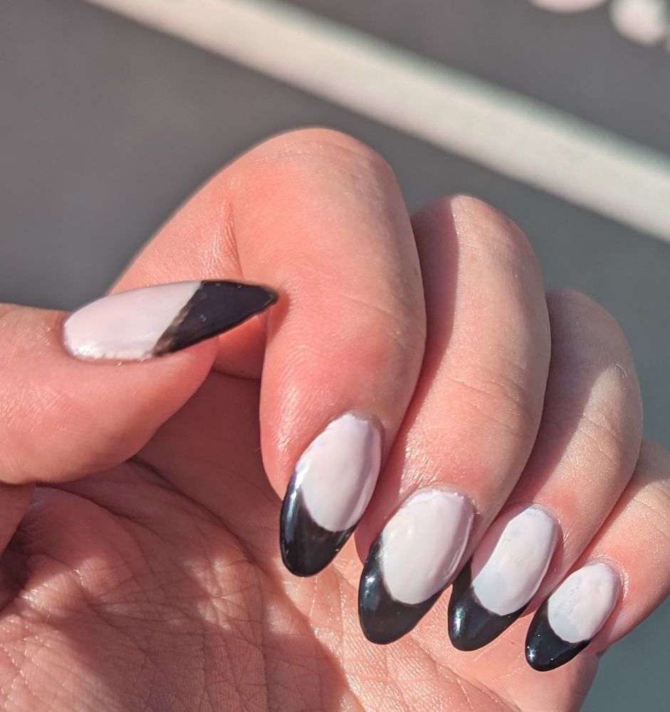 In sunlight, I'm holding my left hand up with fingers curled toward my palm. The nails extend about 6 millimeters past my fingertip, and are almond shaped. They are partially painted with a natural rosy white, which is lighter than my skin tone. The tips are painted with an opaque black in an exaggerated curve, imitating the white tip normally seen on natural nails. The lines where black meets white are slightly messy up close, and the whole nail is slightly rounded outward with a glossy finish. 