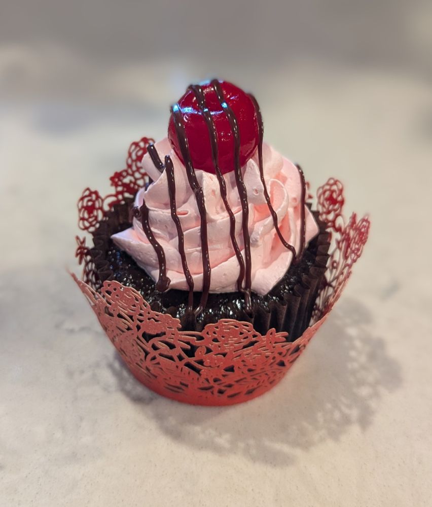 A close up of the cherry chocolate cupcake sitting on a white countertop. The pink frosting is swirled in the center of the dark brown cupcake, with raised ridges. A shiny red cherry with its stem removed sits on top, and a thin line of dark chocolate is drizzled in a zig-zag overtop of the entire thing. Red paper is intricately cut in a lace pattern, and wrapped around the lower part of the cupcake to cover its baking wrapper. 