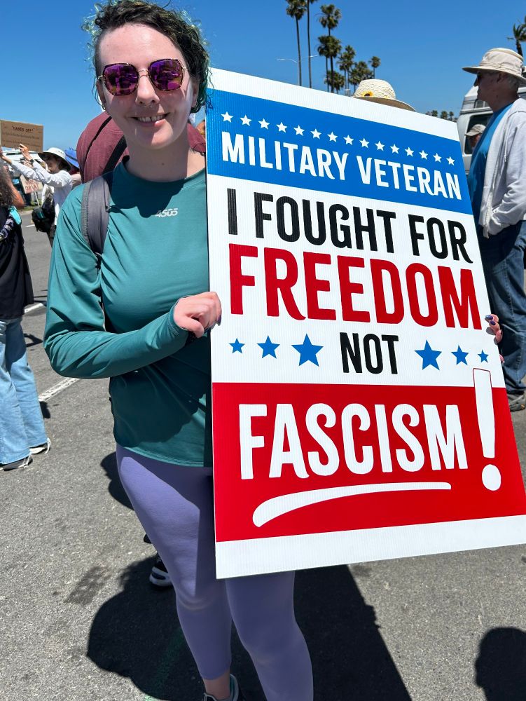 A photo of me under a clear blue sky, in full sunlight on a sidewalk. I'm wearing lavender leggings and a green shirt, holding a 3 foot tall plastic sign. Red and blue text printed on the sign reads, "military veteran. I fought for freedom not fascism!"