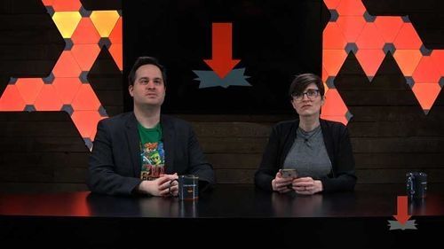 Two people sit at a desk with a bunch of lights and a screen behind them.