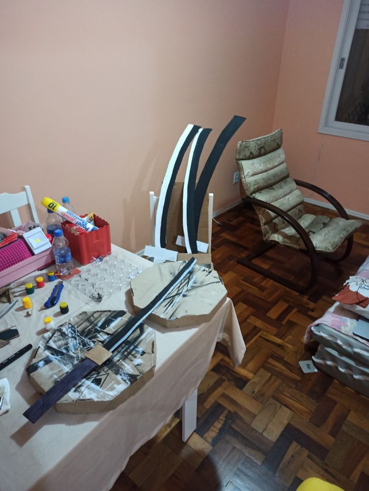 Uma sala cheia de coisas onde o foco é mostrar as katanas de papelão que eu fiz. Tem uma mesa com tintas, estojo e rabiscos e ao redor cadeiras.
