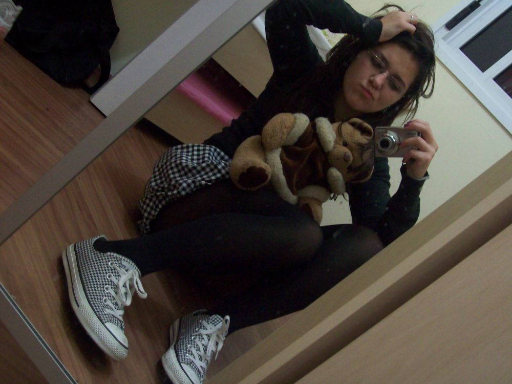 Uma menina fazendo pose e careta, com um ursinho no colo e a mão na cabeça, tirando foto no espelho com a sua câmera digital 