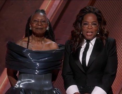 Whoopi and Oprah standing on stage at the 2025 Oscars. Whoopi is wearing a dark blue/gray dress and Oprah is wearing a black suit. 