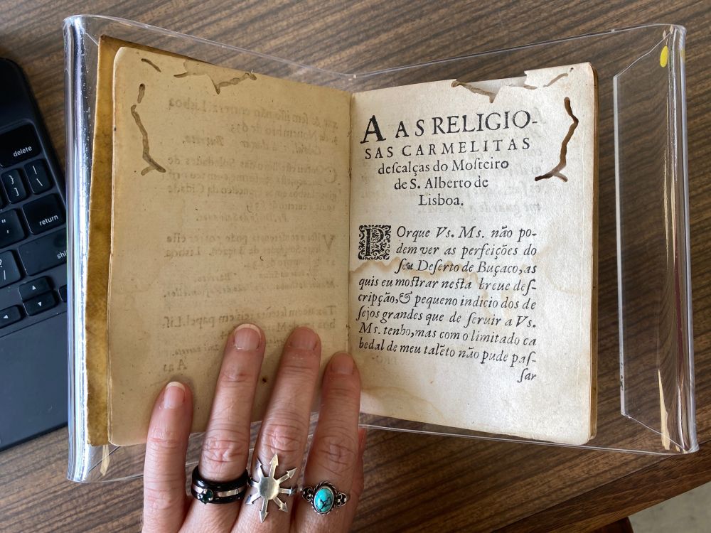 a copy of soledades de buçaco, a 1634 book written by bernarda ferreira de lacerda, rests open on an acrylic book stand, but the blank verso showing is gently held down by my left hand (a white middle aged hand wearing rings on three fingers). the dedication is seen on the recto, with a centered subject line (“To the Discalced Carmelite nuns of the Monastery of Santo Alberto de Lisboa.” – but in Portuguese). The text of the dedication starts with an ornate initial and is set in italic serifed type. The pages have mothwork. There is a formica tabletop underneath (it mimics woodgrain in medium brown shades).