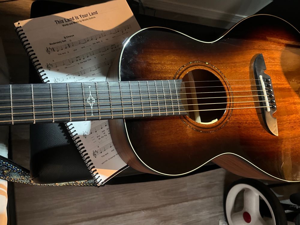 A guitar sits on a bench with music for This Land is Your Land. 