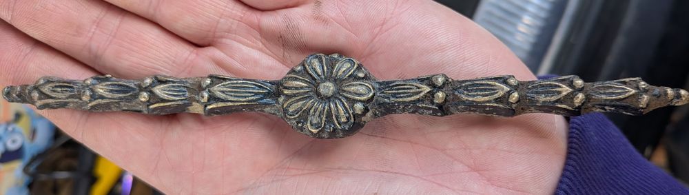 A decorative furniture mount, lying on a human palm. The mount is possibly made from ormolu, and has a central flower with petals, with leaves extending in a linear band to either side. Image credit Glapwell Roots and Remnants