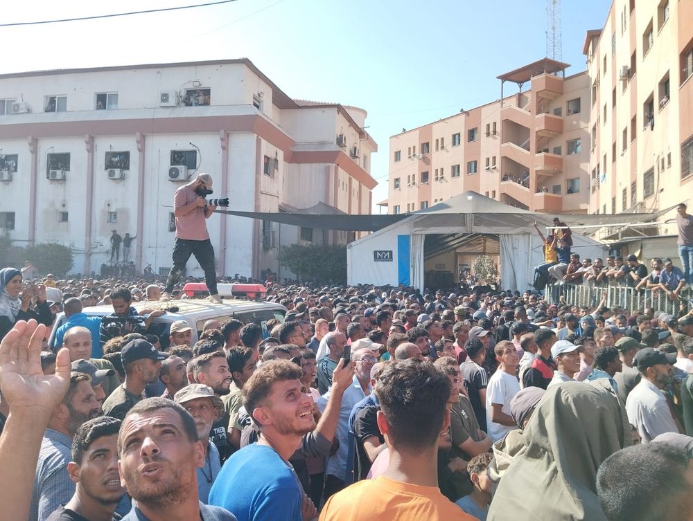 A huge gathering of people, on is standing on a van with a camera in hand.
