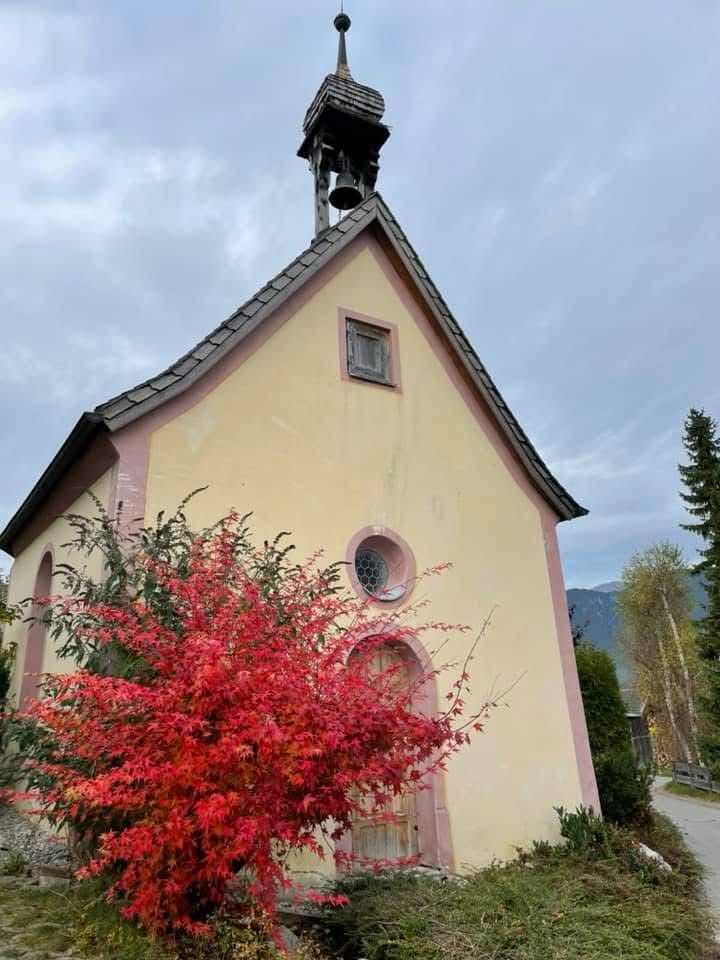 Ein rot gefärbter Busch im Herbst steht vor einer kleinen Kirche mit hellem Mauerwerk und Glockenturm. Das leuchtende Herbstlaub sorgt für einen eindrucksvollen Farbkontrast.