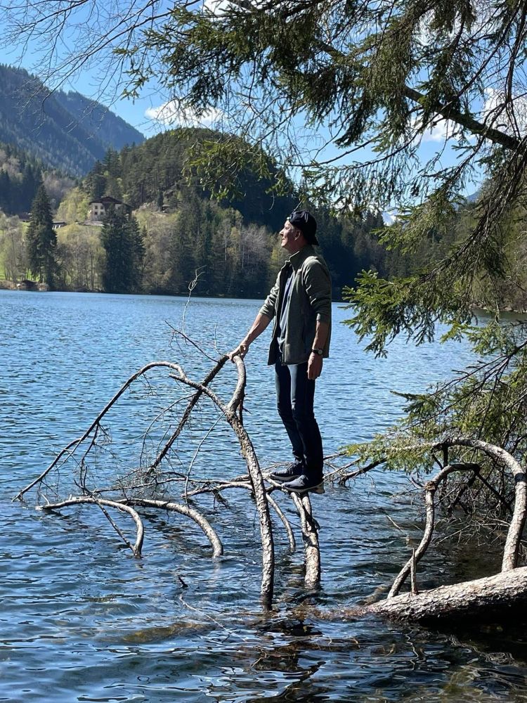 Der Stamm und und Äste eines abgeknickten Baumes liegen im Wasser eines Sees. Auf einem der Äste steht ein Mann, der sich von dort die Gegend anzuschauen scheint.