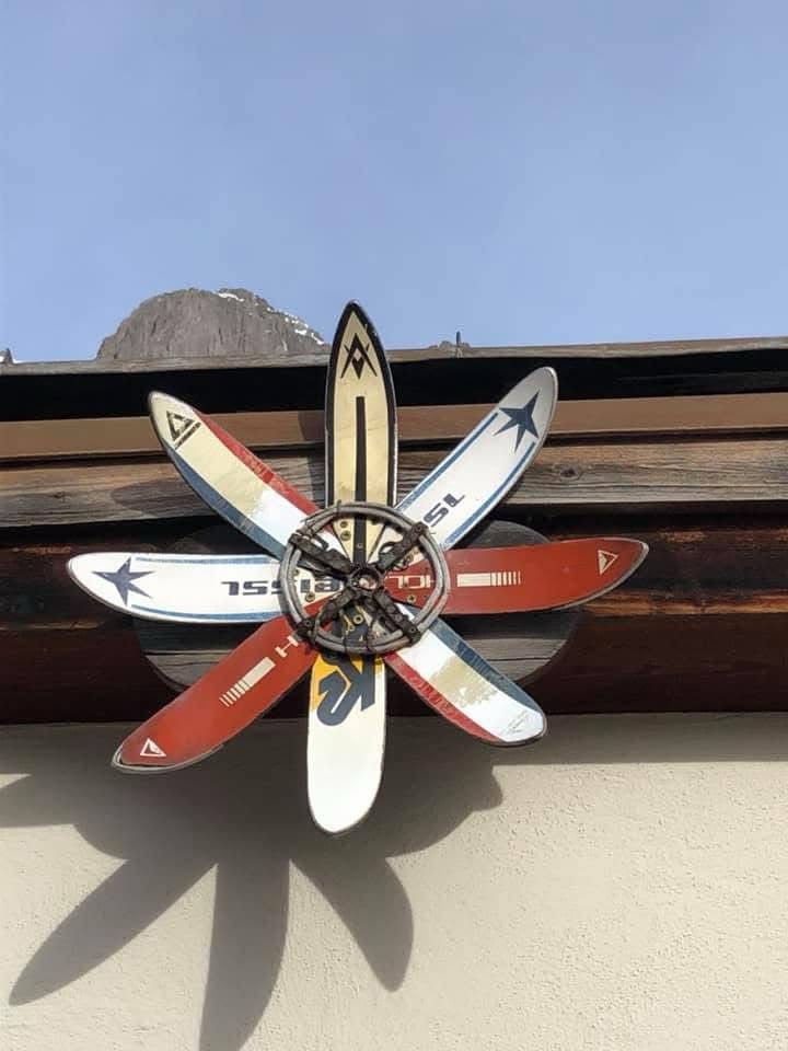 An einem Dach einer Skihütte sind 8 Skispitzen verschiedener Marken in Sternform angebracht! Sie werfen einen Schatten an die Hausmauer! Im Hintergrund ist ein Gipfel erkennbar.