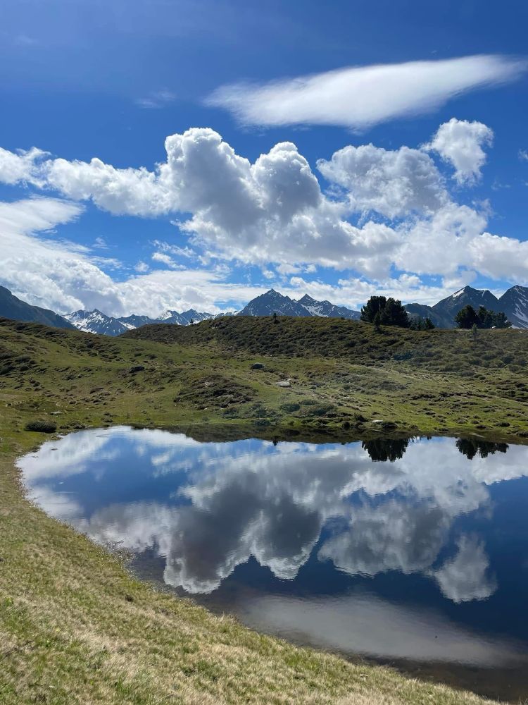 In einem klaren Bergsee spiegeln sich die weißen Wolken und die schneebedeckten Gipfel der umliegenden Berge. Das Bild zeigt die pure Ruhe und Schönheit der alpinen Landschaft unter einem leuchtend blauen Himmel.
